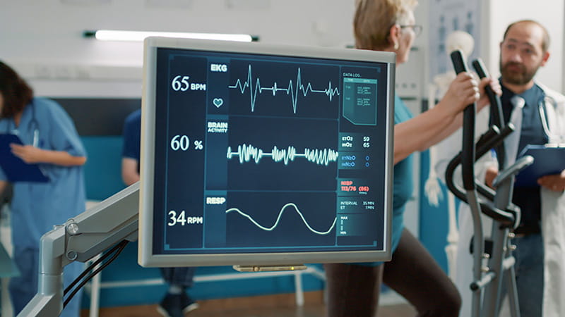 close up of display showing EKG and other medical monitoring information over background of patient using workout equipment while talking to a doctor