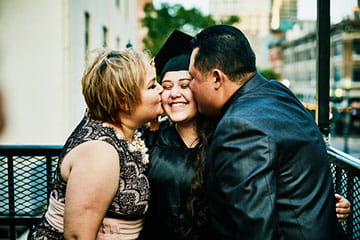 young woman at graduation smiling with her parents kissing her cheeks