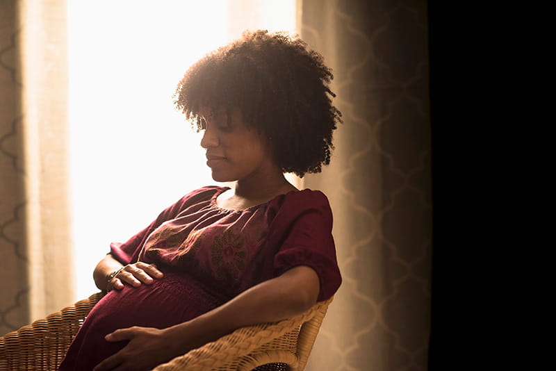 A young pregnant black woman sitting peacefully in a chair, holding her hands on her pregnant belly