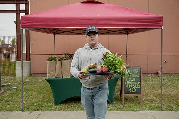 An Hispanic/Latino man is holding a large tray of freshly-picked produce in front of an outdoor Growing Together stand. An Hispanic/Latino man is holding a large tray of freshly-picked produce in front of an outdoor Growing Together stand.
